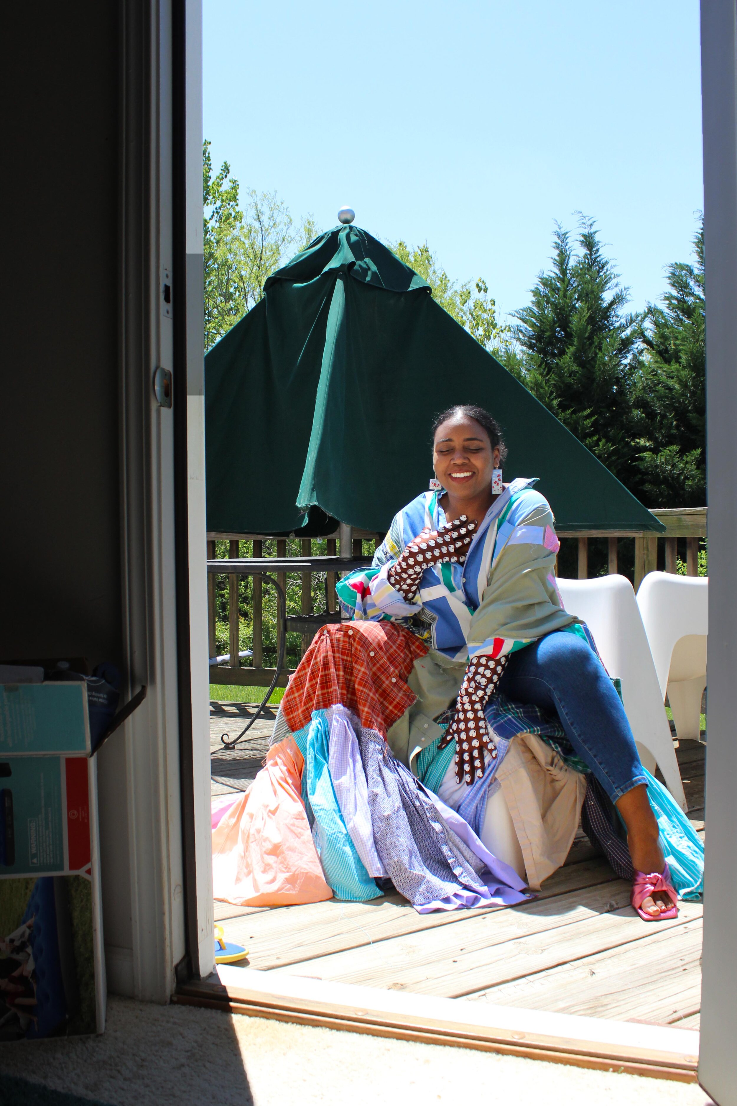 A Black person sits on a white chair on an outside wooden deck, and the photo is shot through a doorway. In the background is another white chair, a green umbrella, and a wooden guard rail, behind that are trees. The model sits with their legs spread and their right arm touches their chest and left arm is down between their legs. The model wears a dress made of multiple squares and patches of fabric. There are patches of green, blue, lavender, green, orange, orange plaid, blue plaid, and white. Underneath are blue cropped skinny jeans and they wear pink open toed heels. Their hair is pulled back and they wear white dangly earrings.