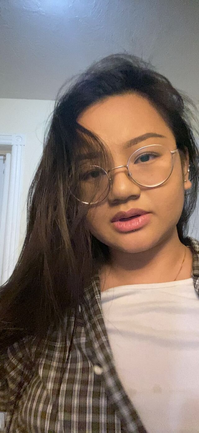 A young person stands with long, black hair, wire-framed glasses. She wears a thin gold chain underneath a white shirt, and a brown and white plaid flannel over it.