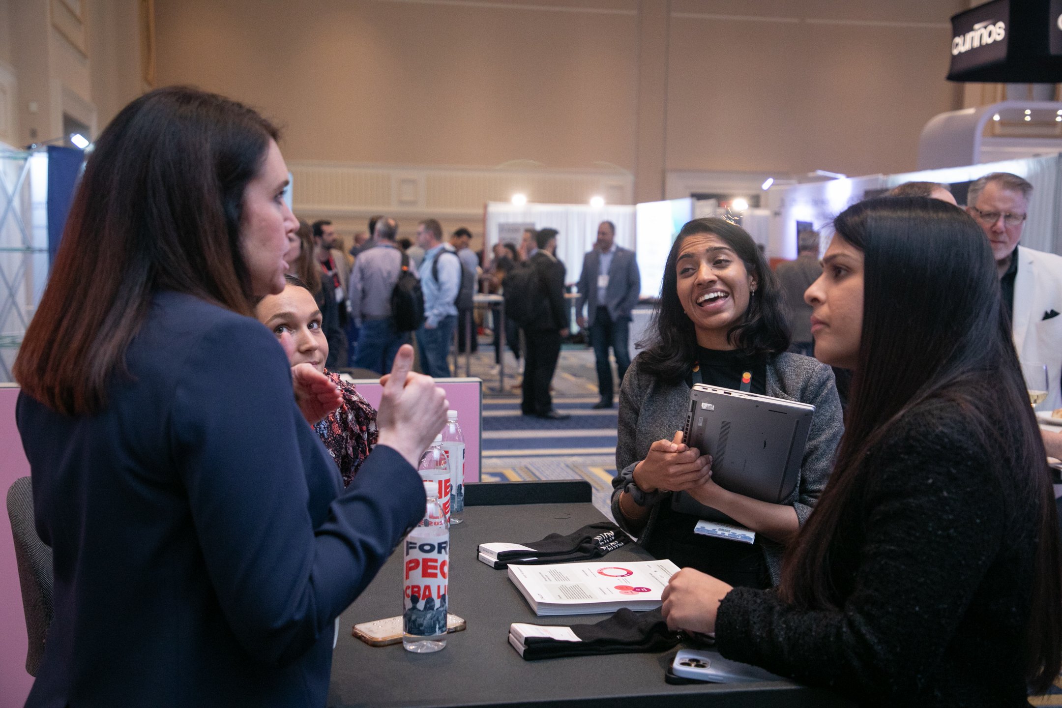 Group of people engaged in conversation at a professional conference or event, standing around a table with books, water bottles, and notepads.